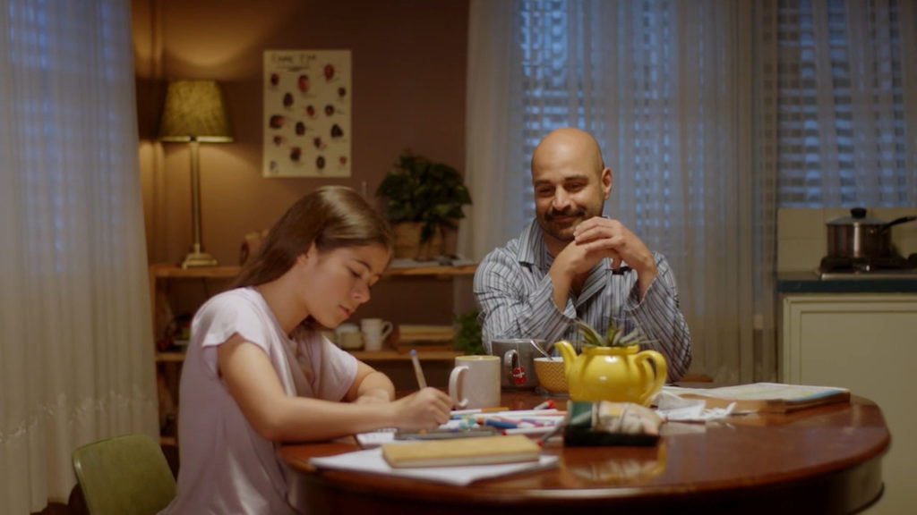A father and daughter sit at a kitchen table together. The father looks on proudly as his daughter does her homework.
